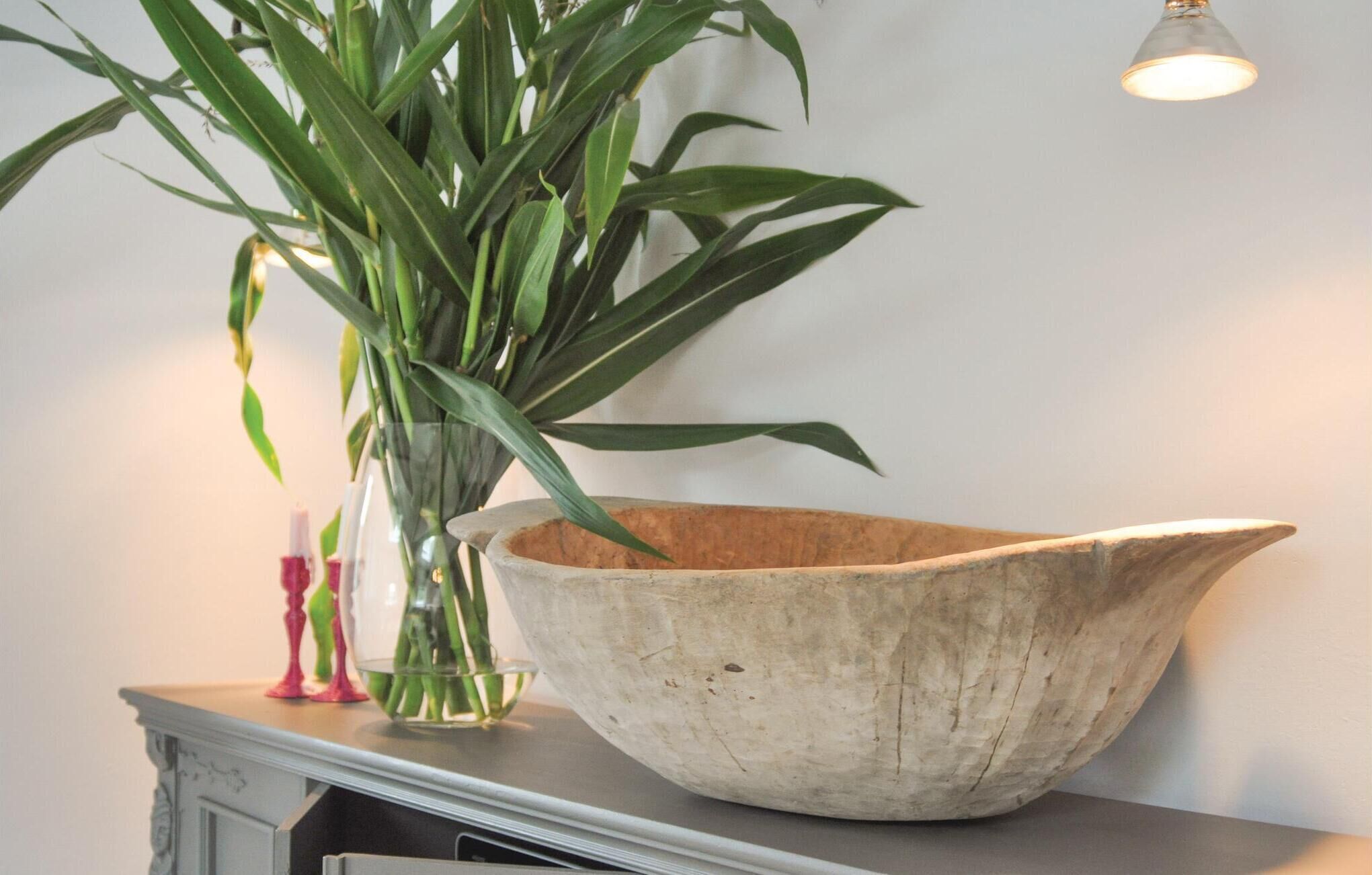 Un grand saladier rustique est posé sur une commode, à côté d’un grand vase transparent contenant une plante verte.