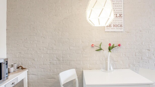 Pared de ladrillo blanco con una mesa y una silla blancas, un jarrón blanco con flores rosadas, y un letrero en la pared con palabras inspiradoras.