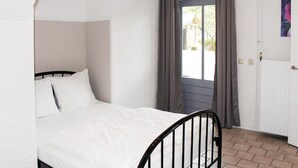 Una habitación pequeña con cama individual, cabecera blanca, ventana con cortinas y puerta con radiador.