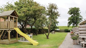 Una estructura de juego de madera con un tobogán amarillo, rodeada de árboles y un césped.