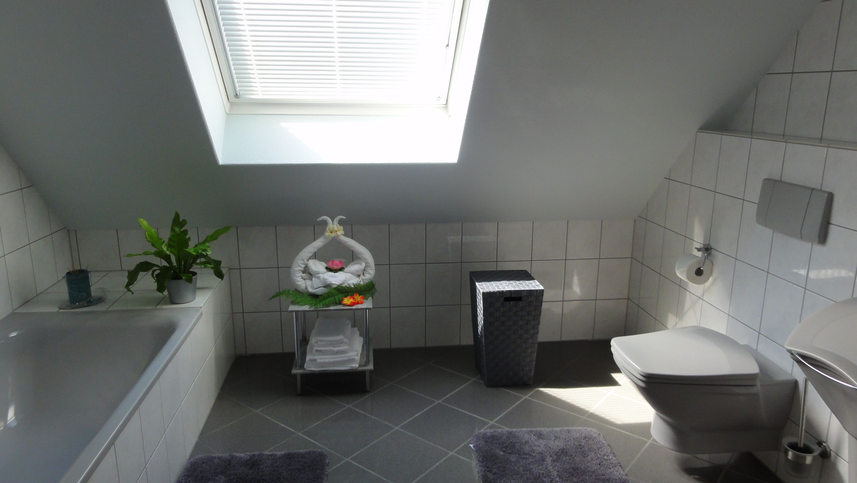 A modern bathroom with a skylight, a bathtub, a toilet, and a small shelf with towels and a potted plant.