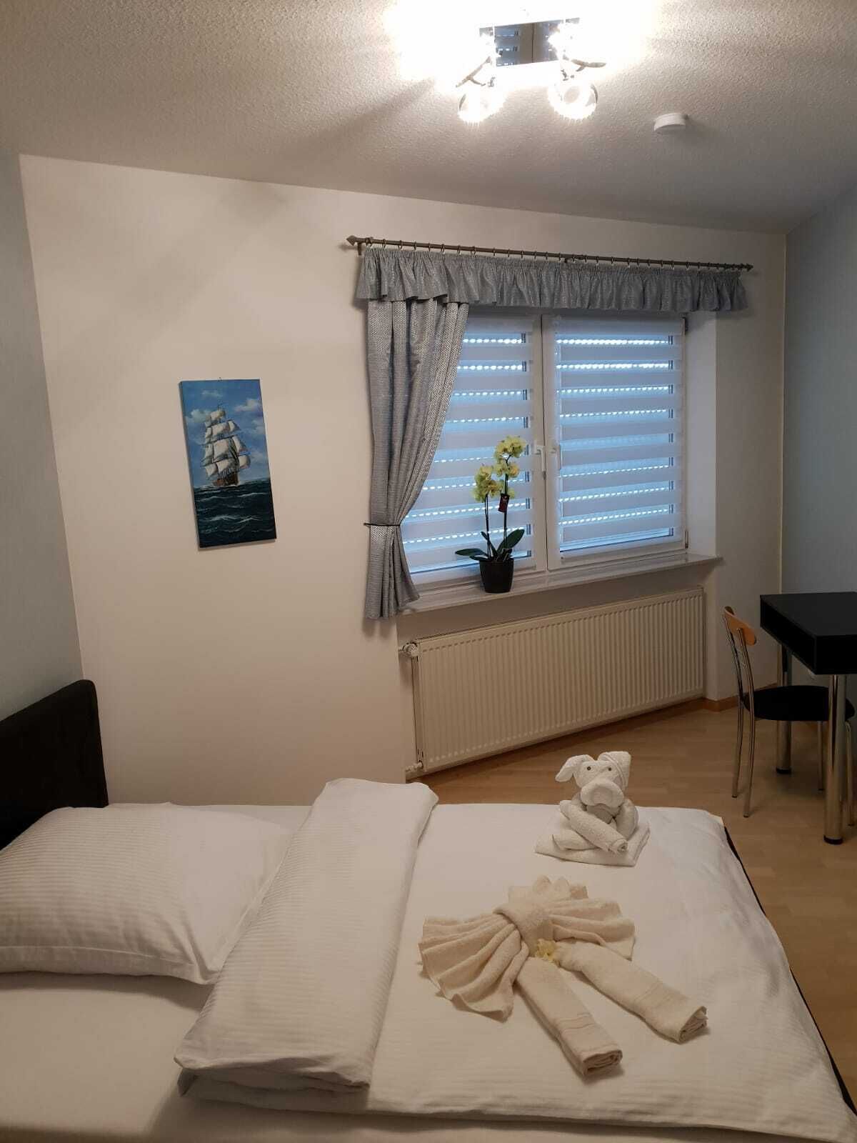 A bedroom with two beds, a window with blinds, a radiator, a small table, and a chair.