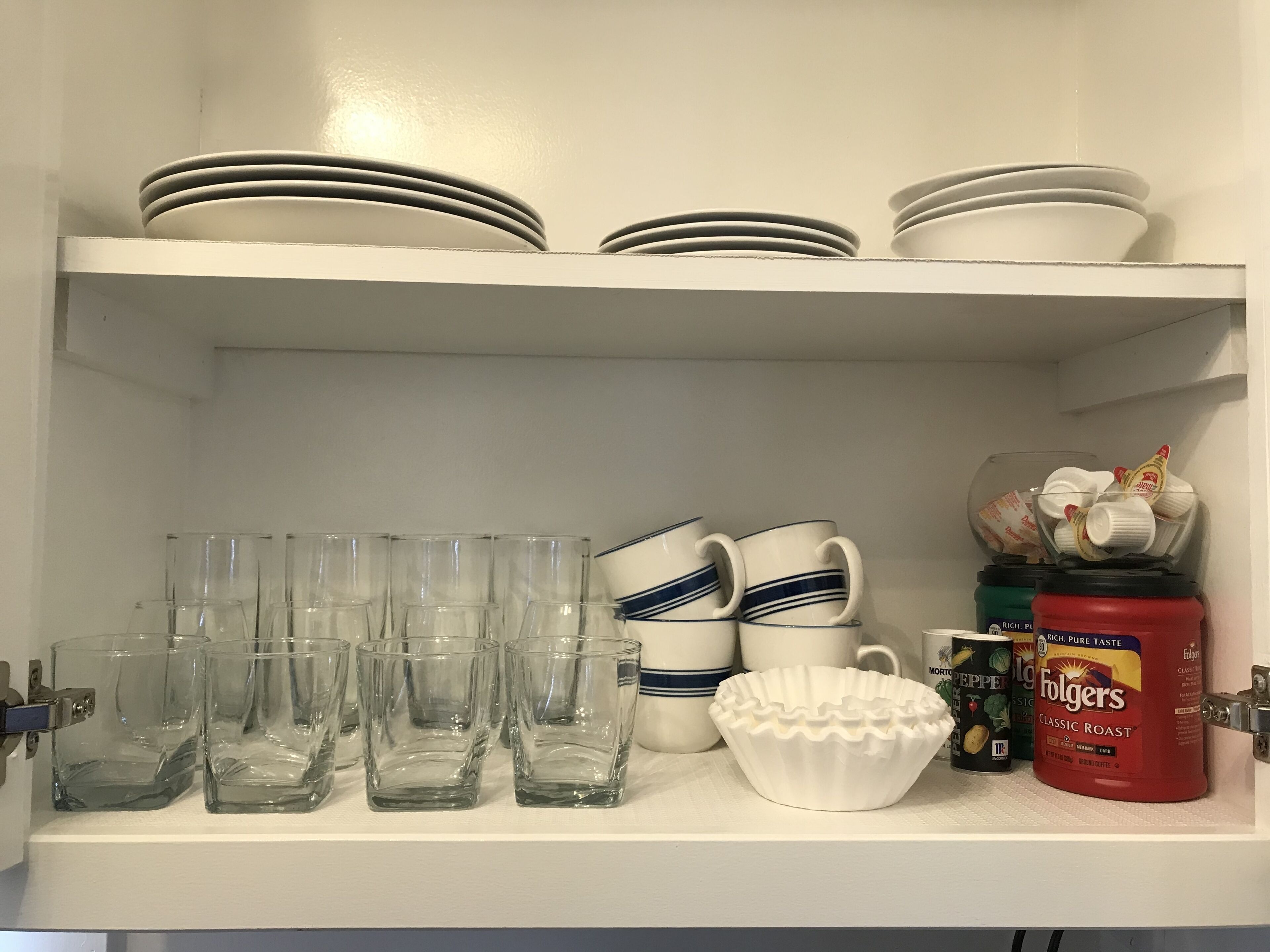 Una estantería en la cocina con platos apilados, vasos transparentes, tazas blancas, un bol blanco y una lata de café Folgers.