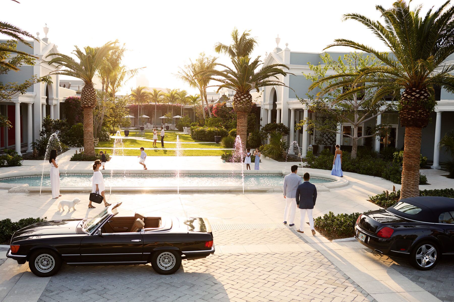 A courtyard with palm trees, a fountain, and a black convertible car parked on the side.