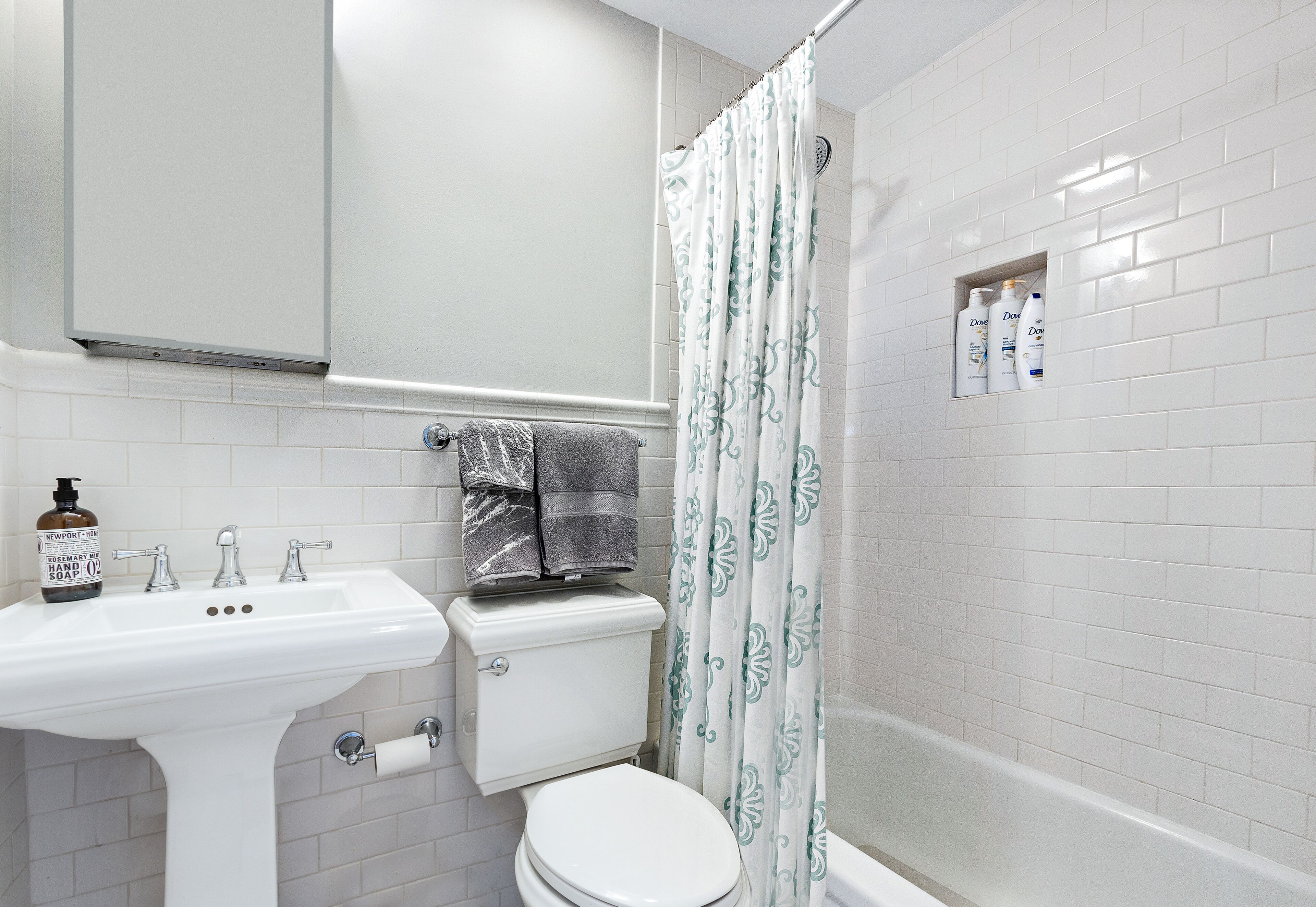 Un baño con lavamanos, inodoro y bañera blancos. Cuenta con una cortina de ducha y un perchero con una toalla.