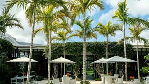 An outdoor patio with white wicker furniture and umbrellas, surrounded by tall palm trees. - Luxury 2 bedroom condo on Palm Beach Island! (Palm Beach)