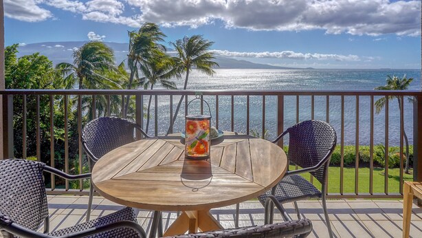 Mesa de madeira redonda com duas cadeiras e um candeeiro decorativo numa varanda com vista para uma praia com palmeiras e para o oceano.
