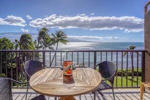 A wooden table with two chairs on a balcony overlooking a beach with palm trees and a view of the ocean.
