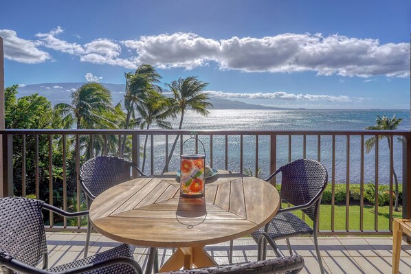 A round wooden table with two chairs and a decorative lantern on a balcony overlooking a beach with palm trees and a view of the ocean.