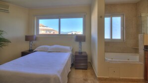 A bedroom with a bed, two bedside lamps, a window with a view, a bathroom with a bathtub, and a mirror. - Farito Beachfront Penthouse, Surf & Paddle Board. (Punta de Mita)