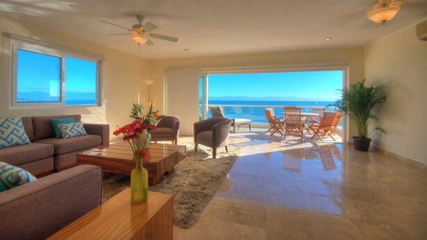 A spacious living room with a large window overlooking the sea, a ceiling fan, and a dining area with outdoor seating. - Farito Beachfront Penthouse, Surf & Paddle Board. (Punta de Mita)