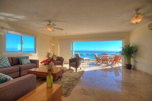 A spacious living room with a large window overlooking the sea, a ceiling fan, and a dining area with outdoor seating.