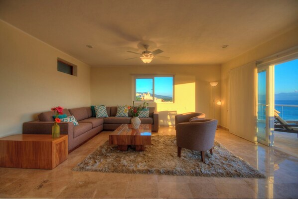 A spacious living room with a large sofa, a coffee table, and a chair. There is a view of the ocean through large windows.
