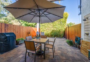 Un patio al aire libre con una mesa y sillas de madera, un paraguas grande y una parrilla de barbacoa.