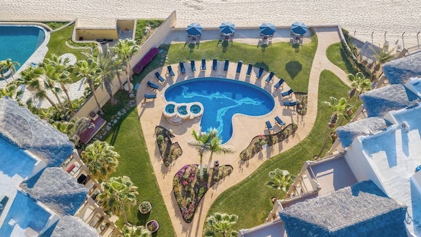 Een resort met een groot rond zwembad, omringd door ligbedden en parasols, een strand op de achtergrond en palmbomen.