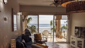 Een woonkamer met een bank, stoelen, een televisie en een balkon met uitzicht op de oceaan.