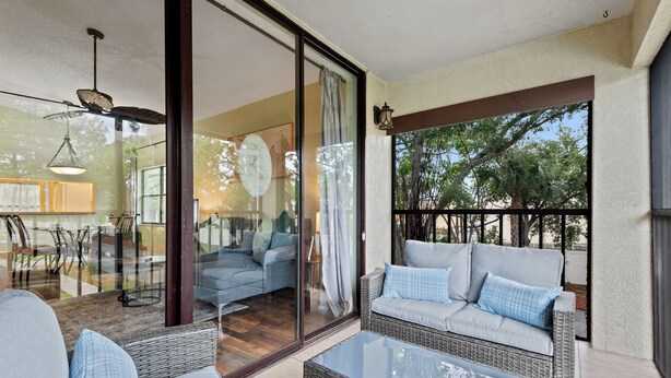 A covered outdoor seating area with wicker furniture and a glass table, overlooking a garden and a building interior. - Stunning & Serene, Minutes to Anna Maria Island, IMG & More (Bradenton)