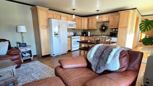 A spacious living room with a brown leather sofa, a wooden coffee table, and a kitchen area with wooden cabinets and appliances. - Lake Front Rental With Chugach Mountain Views! (Glennallen)