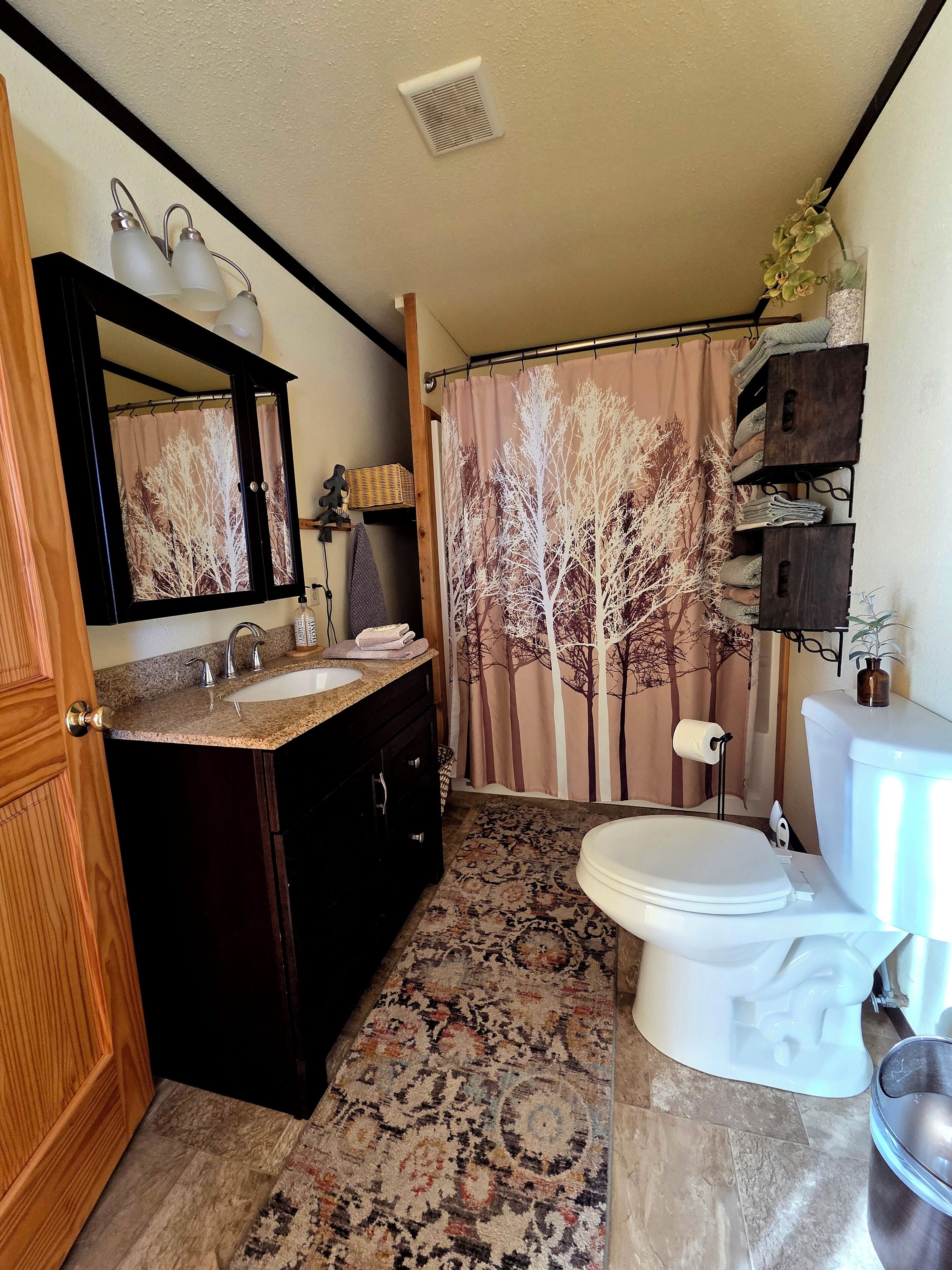 A bathroom with a vanity, sink, mirror, toilet, shower curtain, and a small shelf with towels.