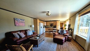 A spacious living room with a brown leather sofa, a wooden coffee table, and a patterned rug. A dining area with a wooden table and chairs is visible in the background. - Lake Front Rental With Chugach Mountain Views! (Glennallen)