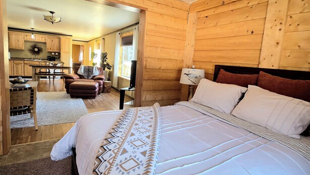 A wooden-paneled bedroom with a large bed, a nightstand, and a view into the kitchen area. - Lake Front Rental With Chugach Mountain Views! (Glennallen)