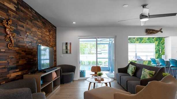 A modern living room with a flat-screen TV, comfortable seating, a wooden accent wall, and large windows offering a view of the outdoors. - ☆Featured on HGTV☆ Heated Pool, SleepNumber Beds, Beach Cruisers (Largo)