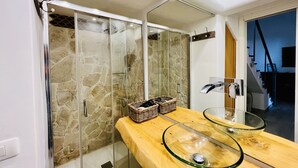 Un baño con ducha de vidrio, tocador de madera y lavamanos de cristal, además de un espejo.