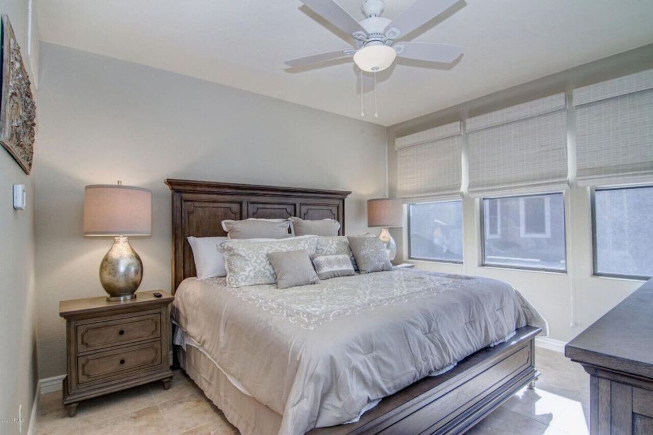 A bedroom with a large bed, nightstands, a ceiling fan, and windows with blinds.