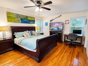 A bedroom with a bed, a desk with a computer, a ceiling fan, and artwork on the walls.