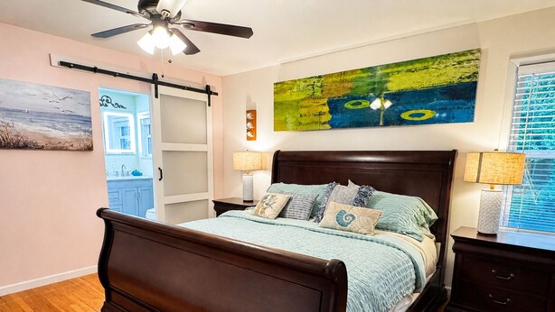 A bedroom with a wooden bed, a ceiling fan, a painting, a window with blinds, and a nightstand with a lamp. - Cheerful Home W/ Private 15' Swim Spa, Big Fenced Yard, & Fire Pit (Arlington)