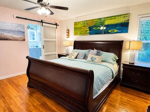 Une chambre à coucher avec un lit en bois, un ventilateur de plafond, un tableau, une fenêtre avec des stores et une table de chevet avec une lampe.