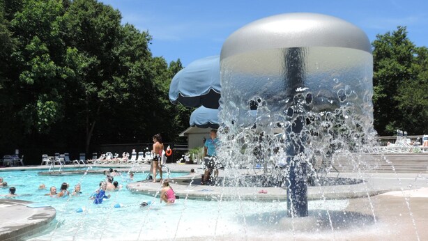 Piscina com escultura de fonte, rodeada por espreguiçadeiras e céu claro.