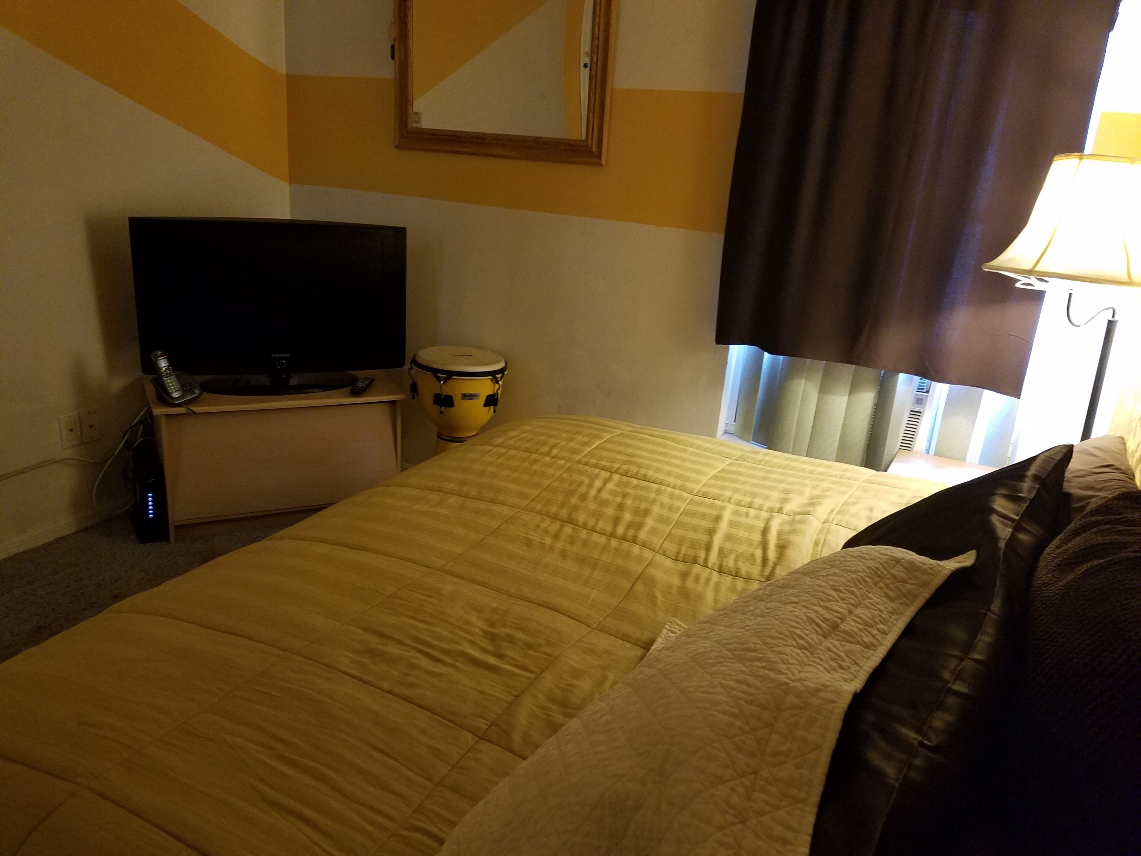 A hotel room with a bed, a television, a drum, and a window with curtains.