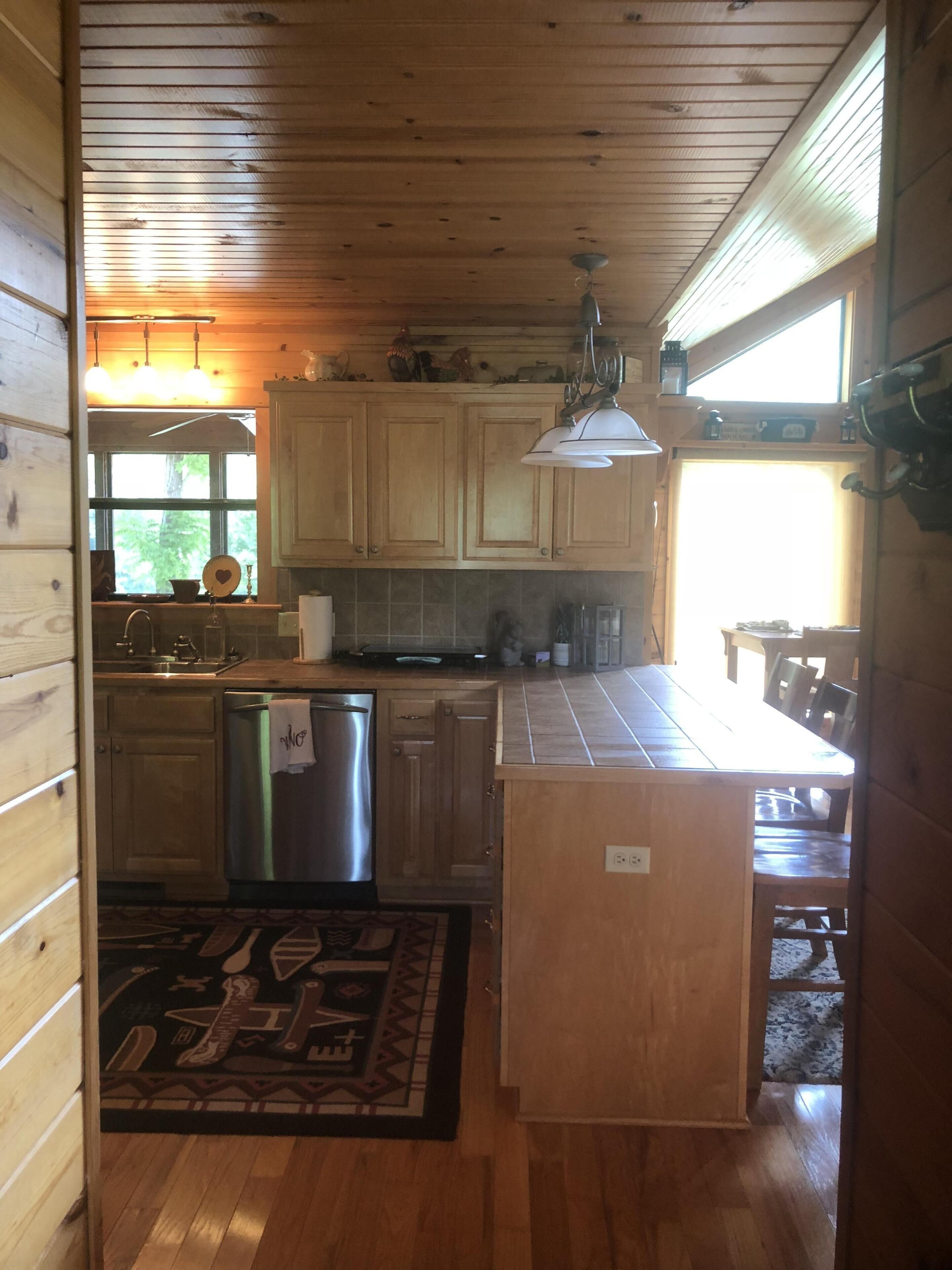 A wooden cabin kitchen with a central island, wooden cabinets, a stainless steel dishwasher, and a dining area with a table and chairs.