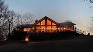 Une grande cabane en bois à deux étages, dotée d’une cheminée centrale imposante, et entourée d’une cour clôturée.