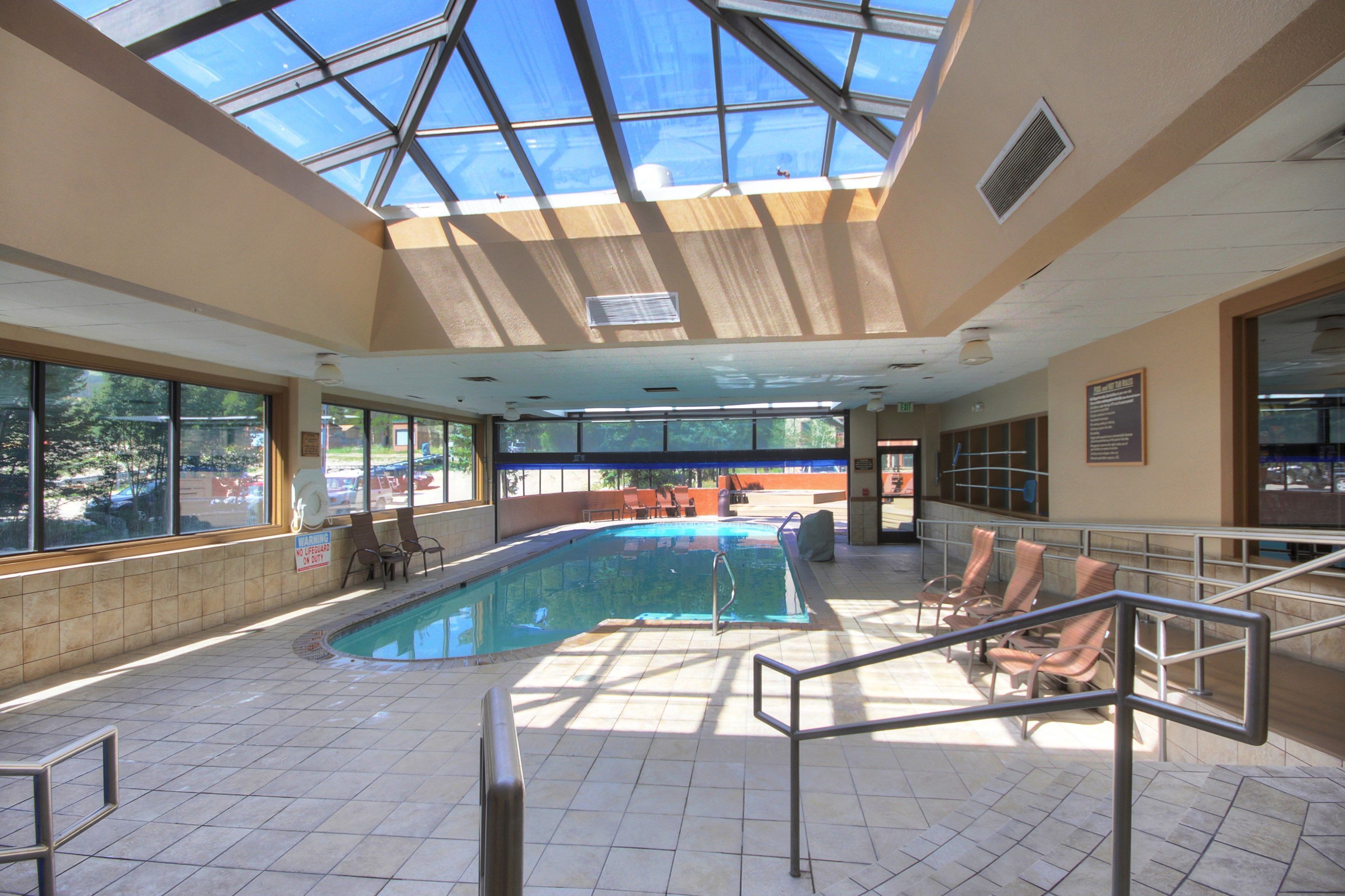 An indoor swimming pool with a glass ceiling, sunlit area, and seating by the pool.
