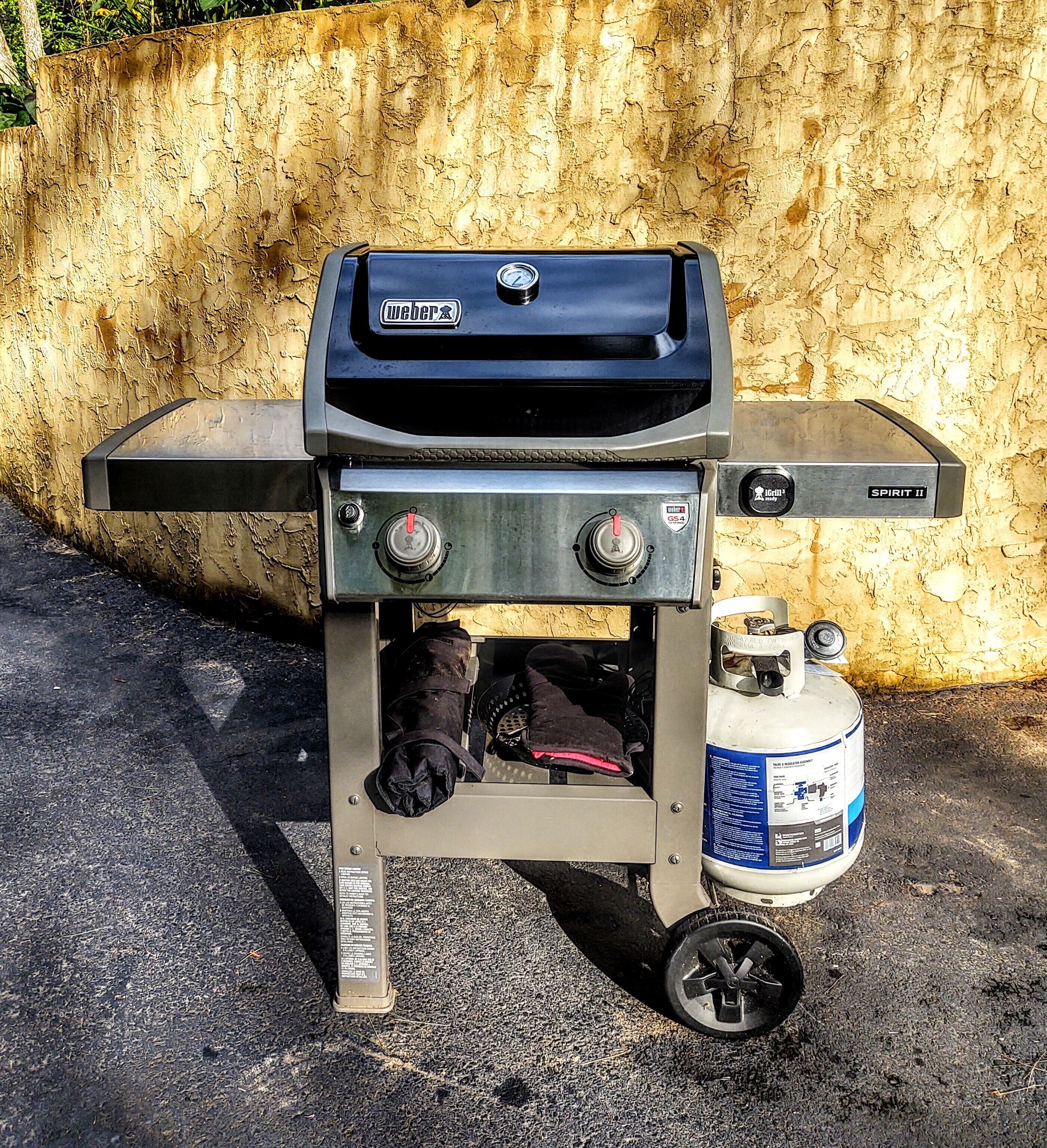 Een draagbare Weber Spirit II grill met een bijzettafeltje, een gasfles en een paar wanten die aan de zijkant hangen.