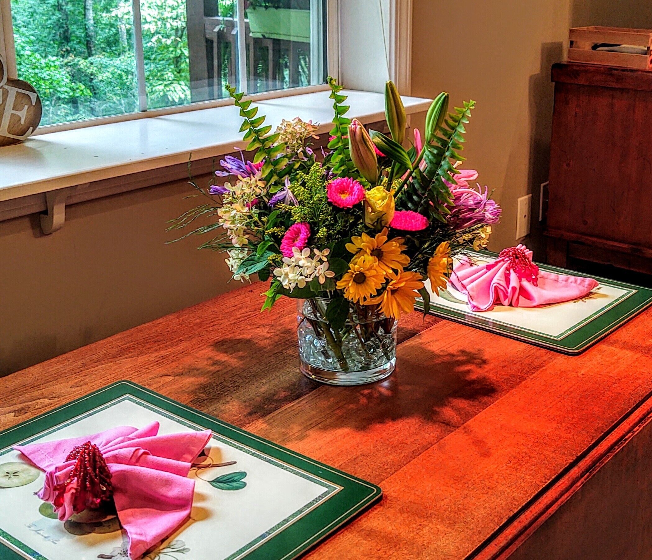Una mesa con un centro de mesa floral, un armario de madera y una ventana con vistas a follaje.
