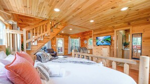 A wooden cabin interior with a bed, TV, staircase, and a view of the outdoors.