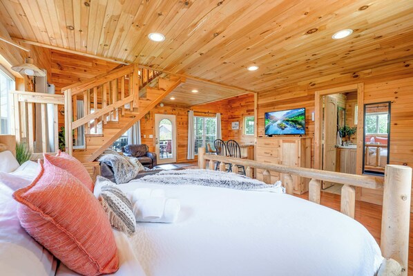 Interior de una cabaña de madera con cama, televisor, escalera y vistas al exterior.
