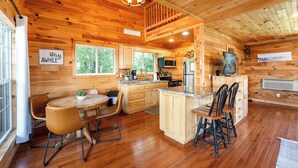 A wooden cabin interior with a kitchen, dining area, and a large window.