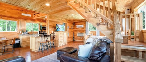 A wooden cabin interior with a living area, kitchen, and a staircase.