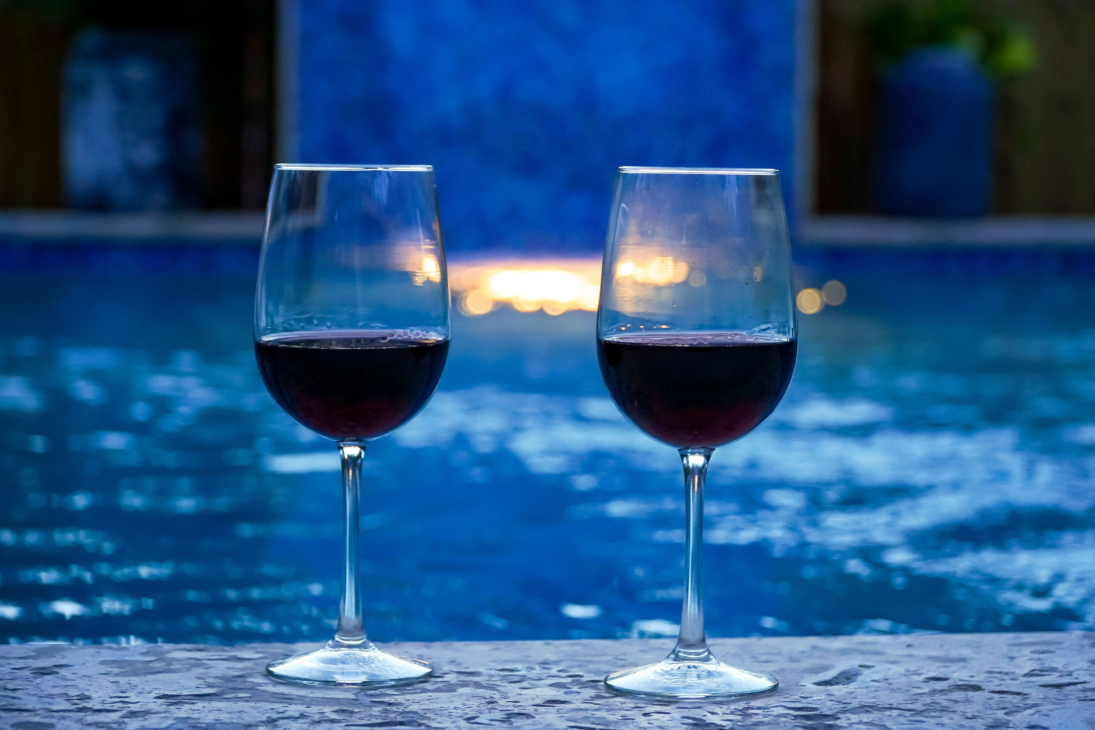 Two glasses of red wine on a table by a pool with a sunset in the background.