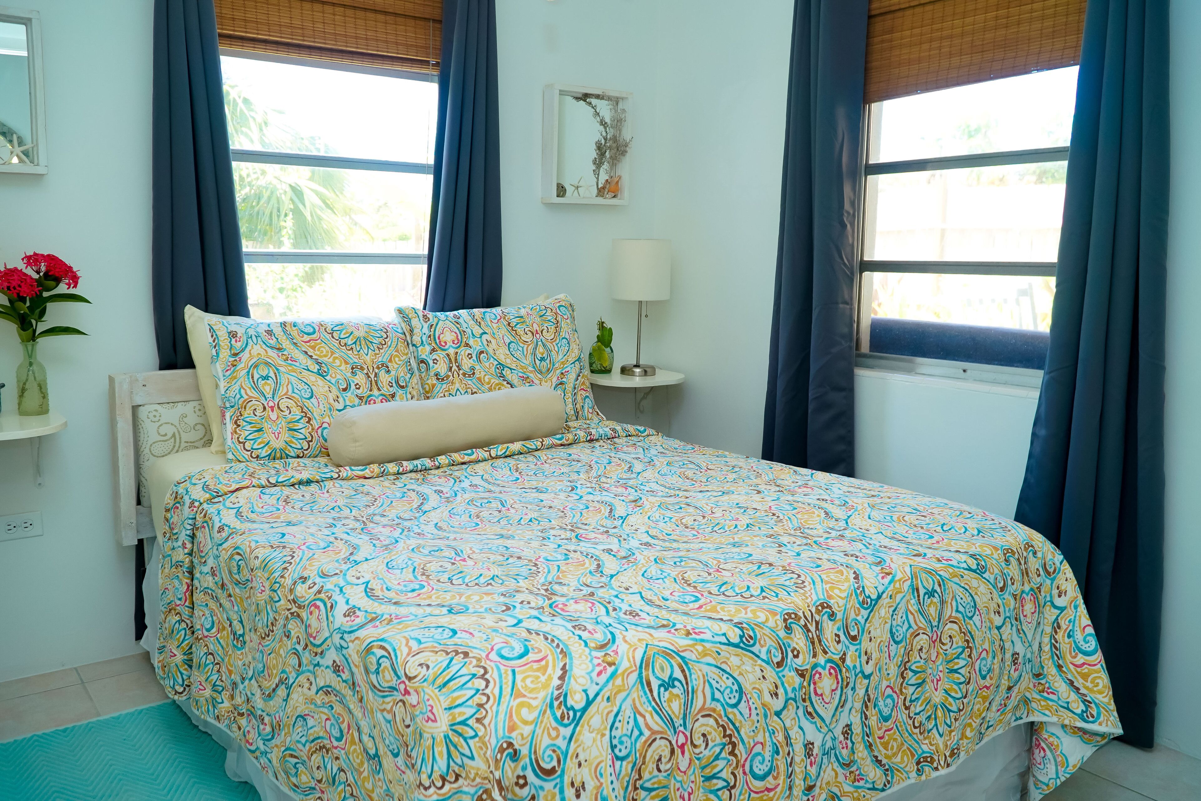 Une chambre à coucher avec une couvre-lit à motifs, une table de chevet avec une lampe et une fenêtre avec des rideaux.