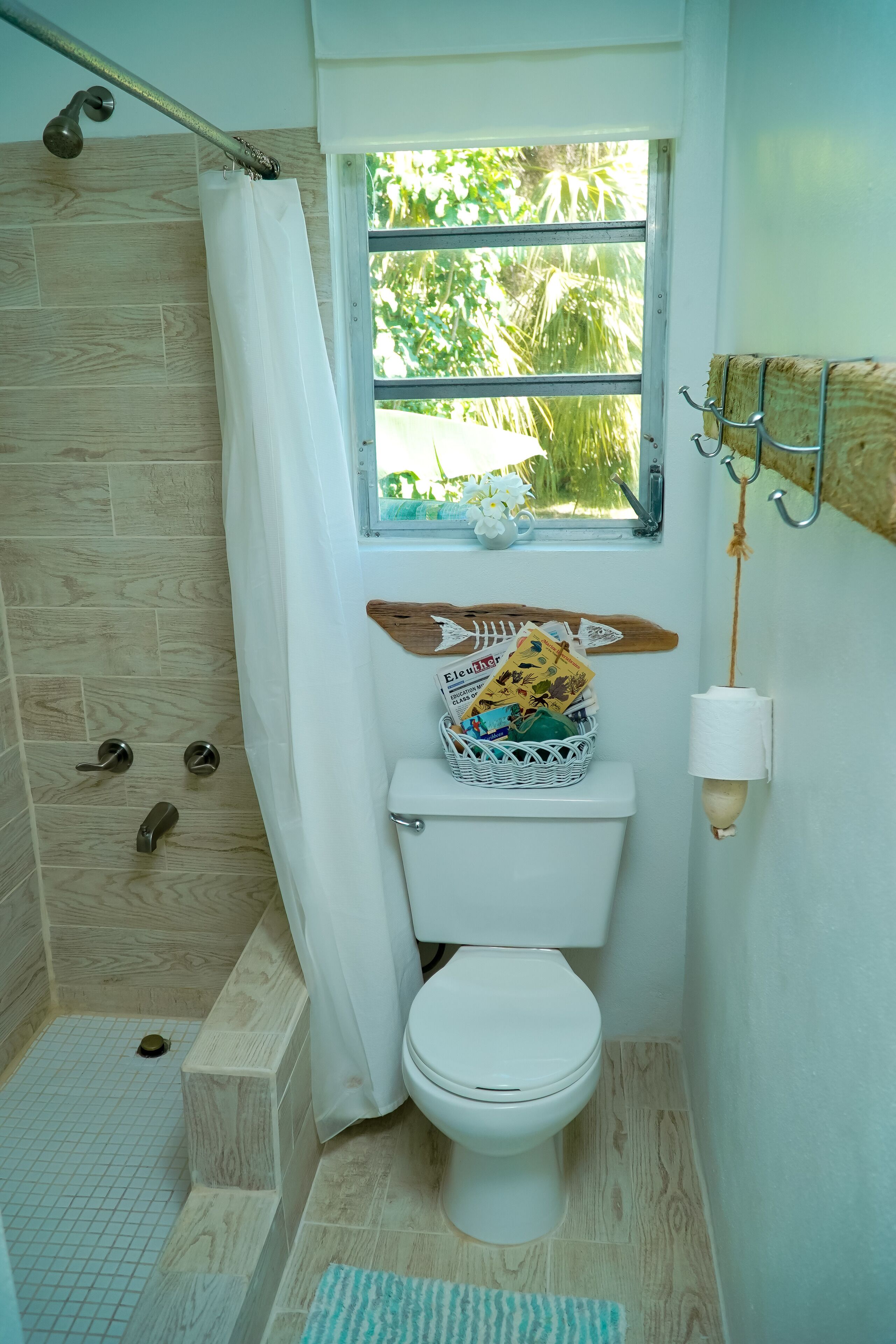 A compact bathroom with a shower, a window with a view of greenery, a white toilet, and a basket of toiletries.