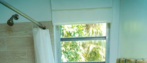 A compact bathroom with a shower, a window with a view of greenery, a white toilet, and a basket of toiletries.