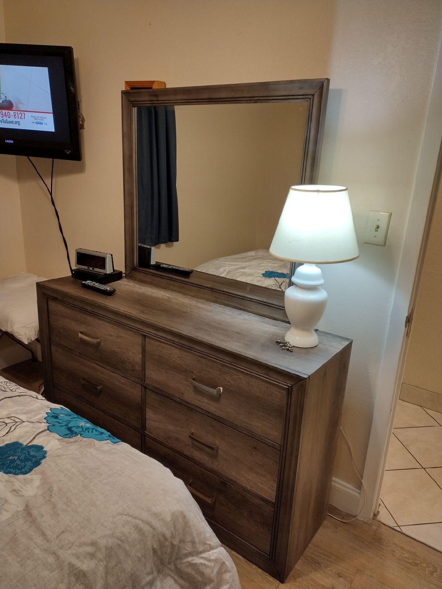 A wooden dresser with drawers, a mirror, a lamp, and a television mounted on the wall.