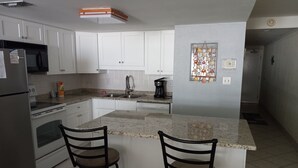A kitchen with white cabinets, a stainless steel refrigerator, a microwave, a coffee maker, and two bar stools.