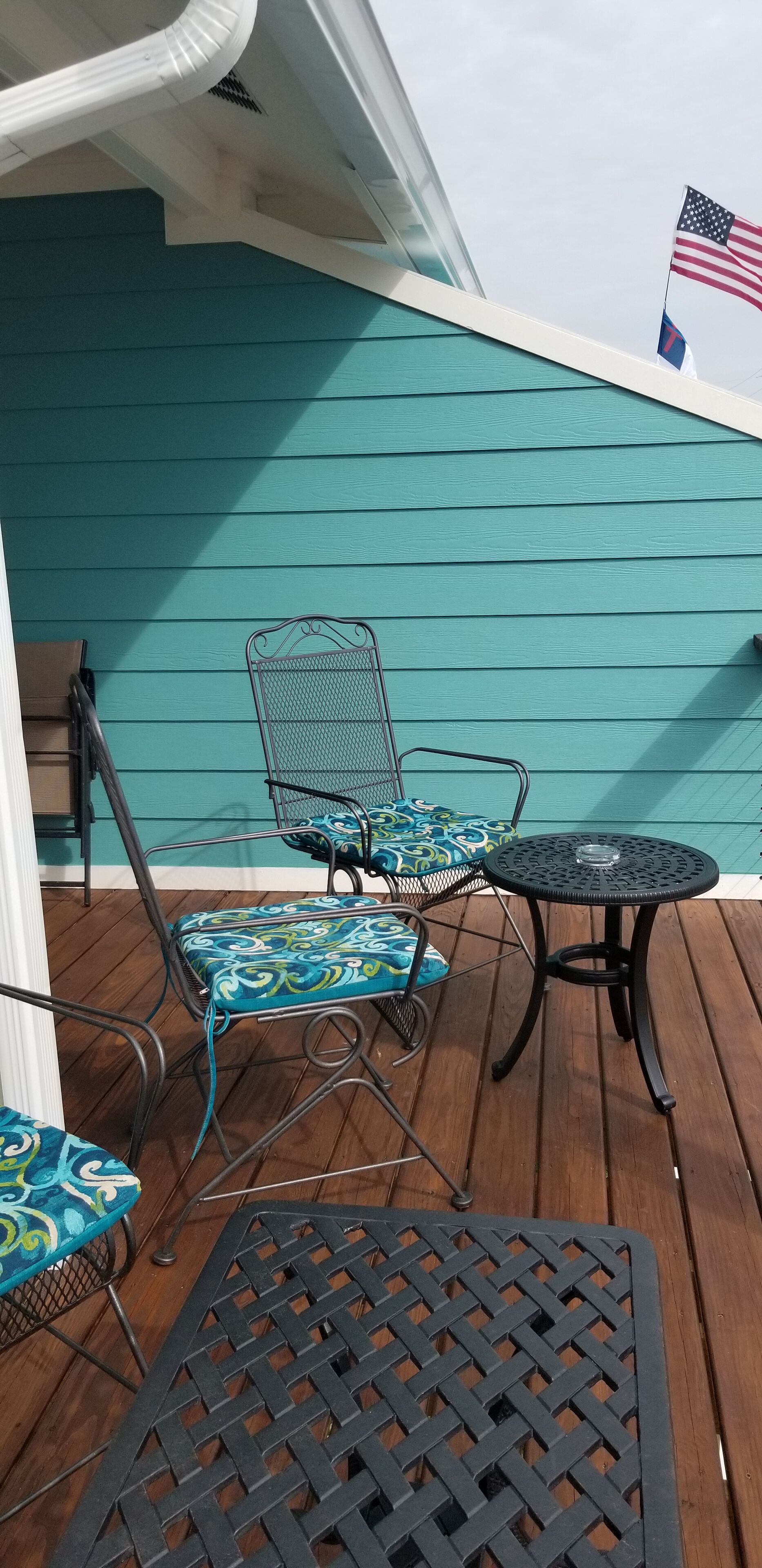 Un espace extérieur abrité, avec des murs de couleur turquoise, une table en métal et deux chaises dotées de coussins à motifs.