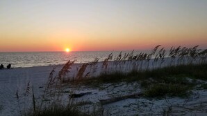 Ein Strand bei Sonnenuntergang mit Sand und Gras im Vordergrund, die Sonne geht über dem Wasser unter.